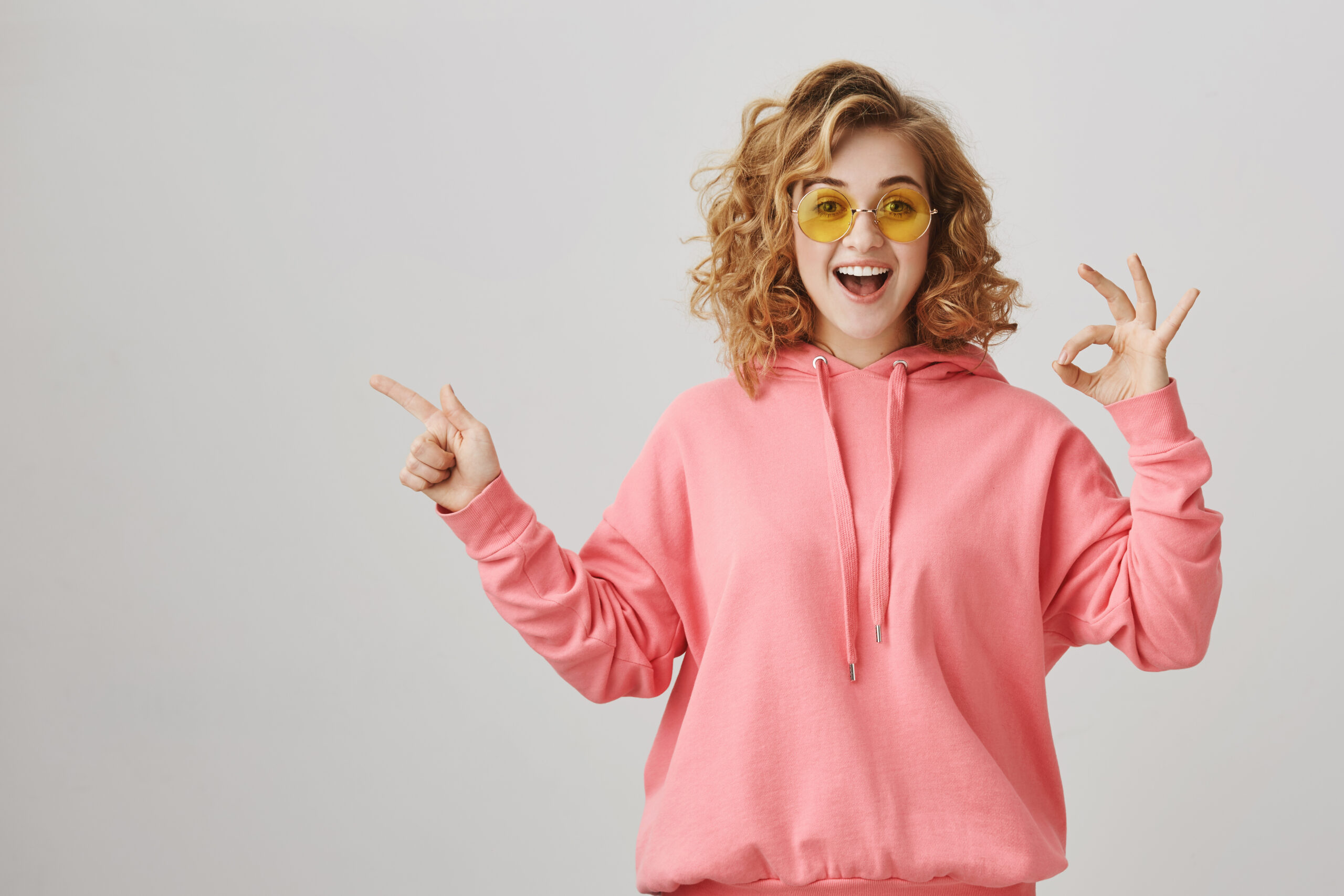 Studio portrait of emotive happy feminine woman with curly hair, wearing casual hoodie and sunglasses showing ok sign and pointing left with index finger, smiling with excitement over gray background